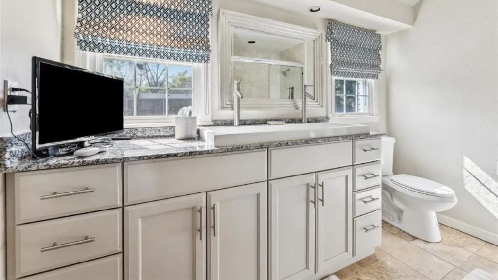 A peaceful bathroom with a vanity, large mirror, white toilet, and windows with blue curtains.
