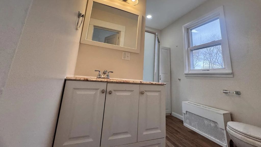 A cozy bathroom with wooden floors, a granite vanity, large window, and glass shower.