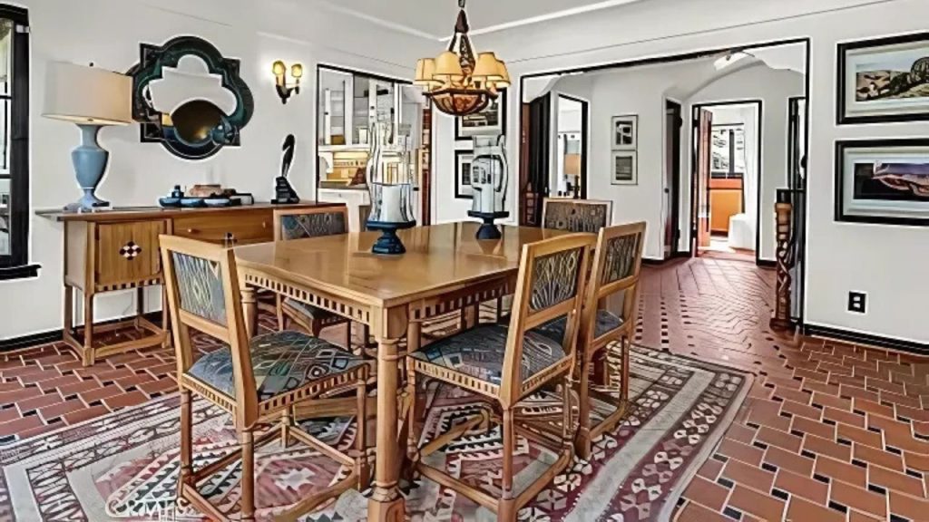 A cozy dining area with a wooden table, terracotta tiles, a chandelier, and framed artwork, opening to other rooms.