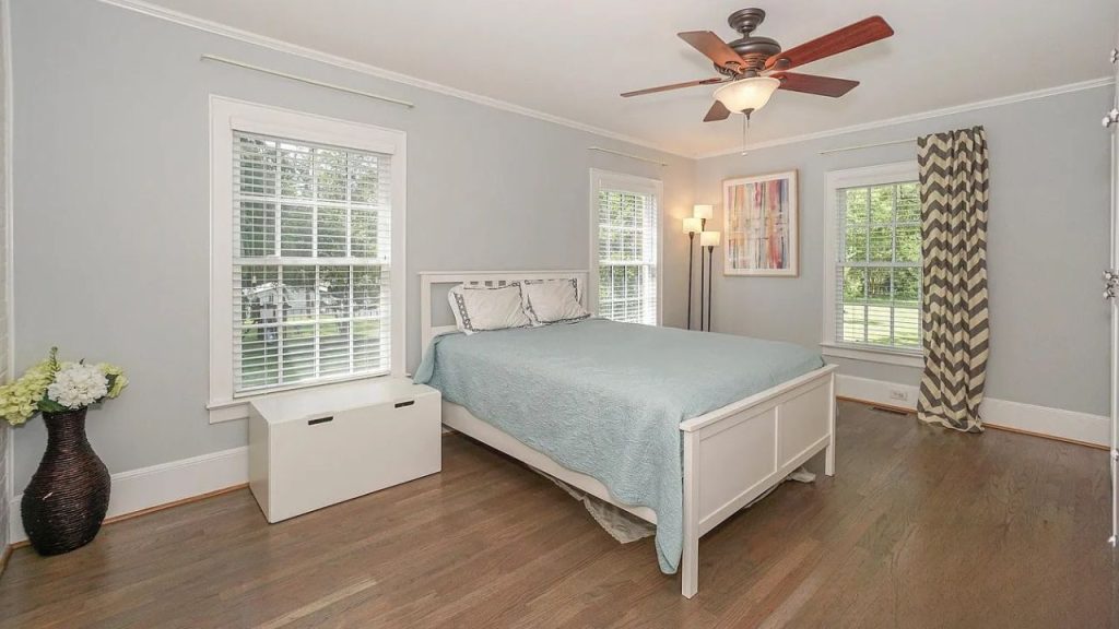 A serene bedroom with a teal quilted bed, large windows, a ceiling fan, and warm hardwood floors.