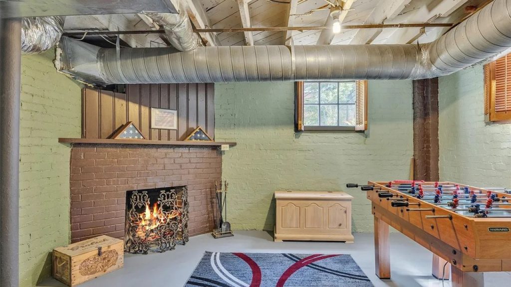 A cozy game room with a brick fireplace, foosball table, green walls, and exposed ductwork in a basement setting.