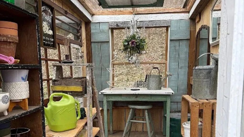 A charming garden shed with a wooden table, gardening tools, a chandelier, and green walls under a glass ceiling.