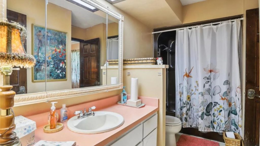 A serene bathroom with a pink countertop, single sink, floral shower curtain, and skylight, featuring a vintage lamp and wicker baskets.