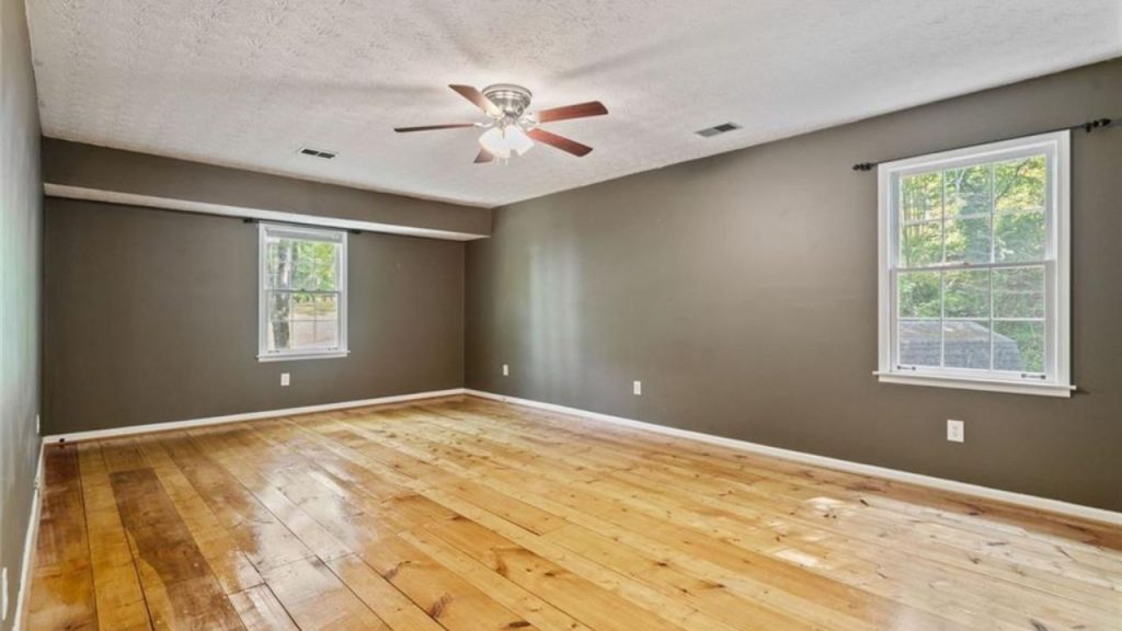 A spacious bedroom with gray walls, wooden floors, large windows, and a ceiling fan.