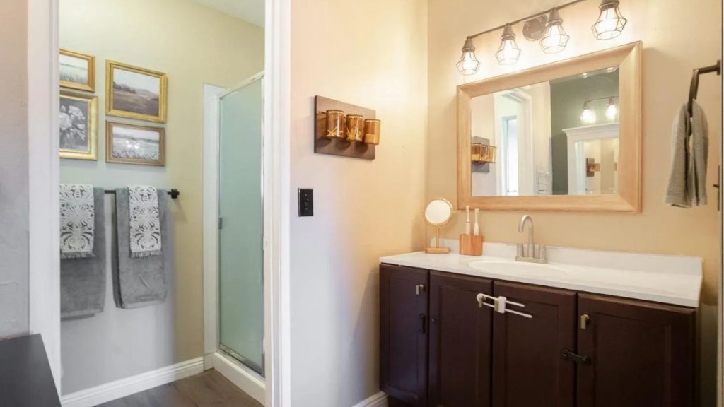 A cozy bathroom with a glass shower, dark vanity, round mirror, amber glass holders, and patterned towels under warm lighting.