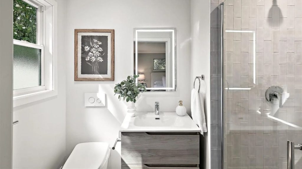 A bright bathroom with a backlit mirror, frosted window, and glass shower, accented by plants and artwork.
