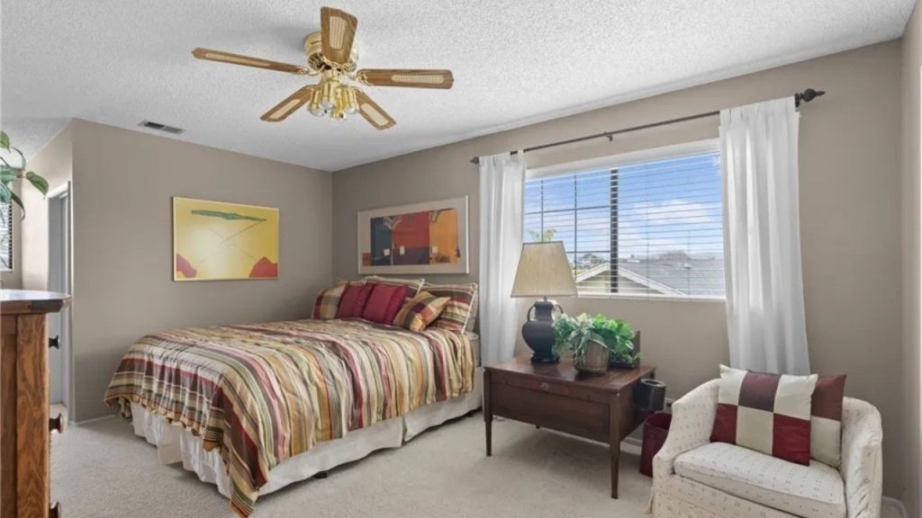 A cozy guest bedroom with a striped bed, wooden nightstand, large windows, and a comfy armchair.