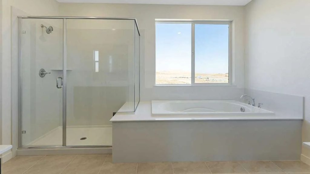 A serene bathroom with a large soaking tub by a window, a glass-enclosed shower, beige tiles, cream walls, and a spacious countertop under soft natural light.