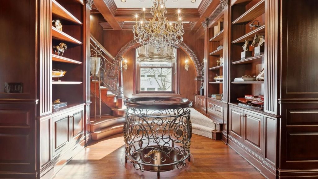 A sophisticated study with mahogany bookshelves, a chandelier, a wrought-iron table, and a curved staircase, featuring a large window with ornate woodwork and hardwood floors.
