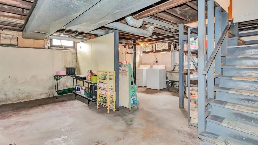 A rustic basement with exposed pipes, a washer and dryer, metal shelves with supplies, a concrete floor, and a weathered staircase leading upstairs.