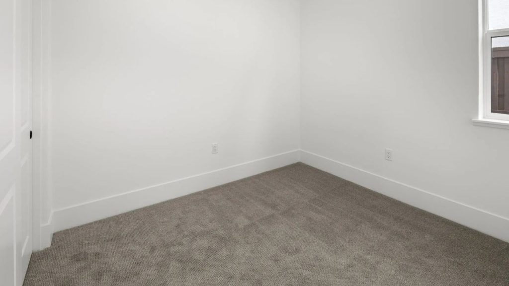 A small bedroom with gray carpeting, white walls, and a window overlooking a fenced backyard.