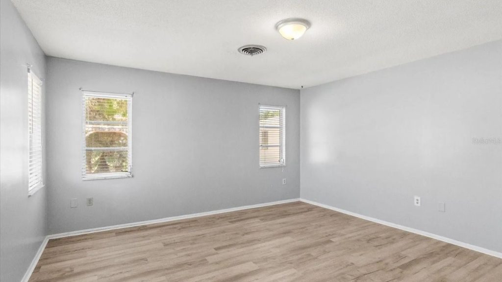A spacious bedroom with light gray walls, light wood floors, two windows with white shutters, and a simple ceiling light fixture.