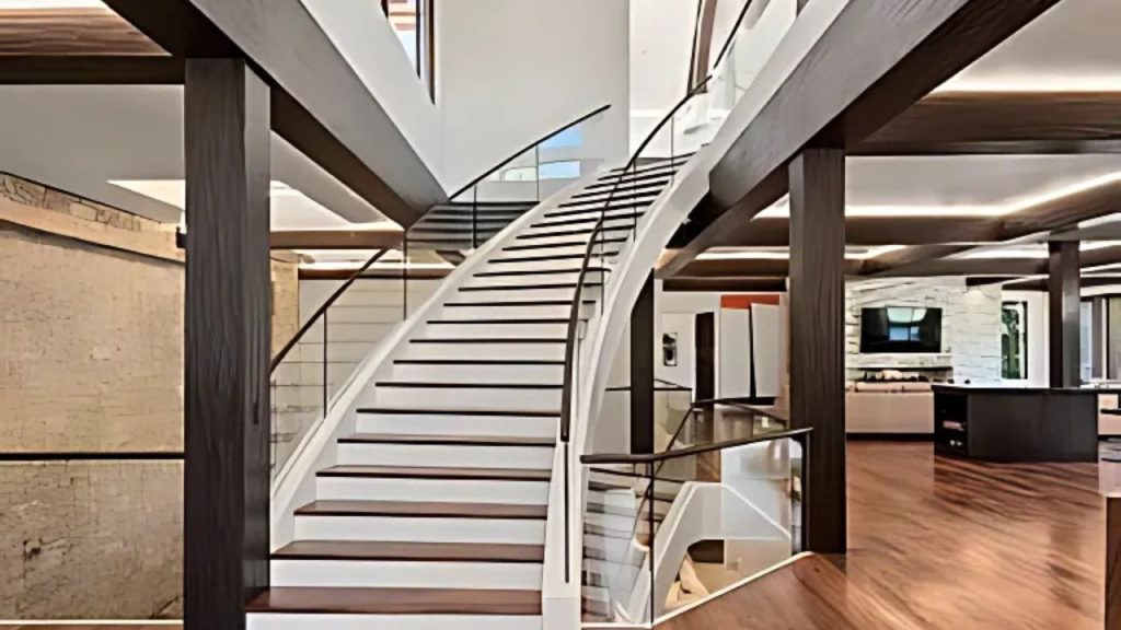 A grand staircase with glass railings and wooden beams, leading to an open area with wooden floors and a kitchen island in view.