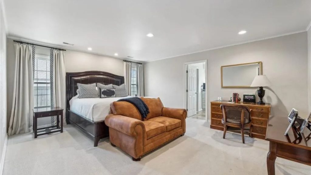 A cozy bedroom with a brown sofa, dark wood bed, desk, and large windows with curtains.