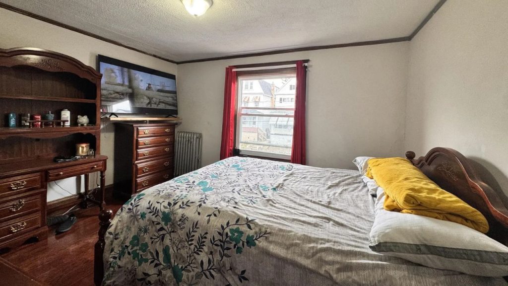 A cozy bedroom with wooden floors, a patterned bed, red-curtained window, and wooden furniture.