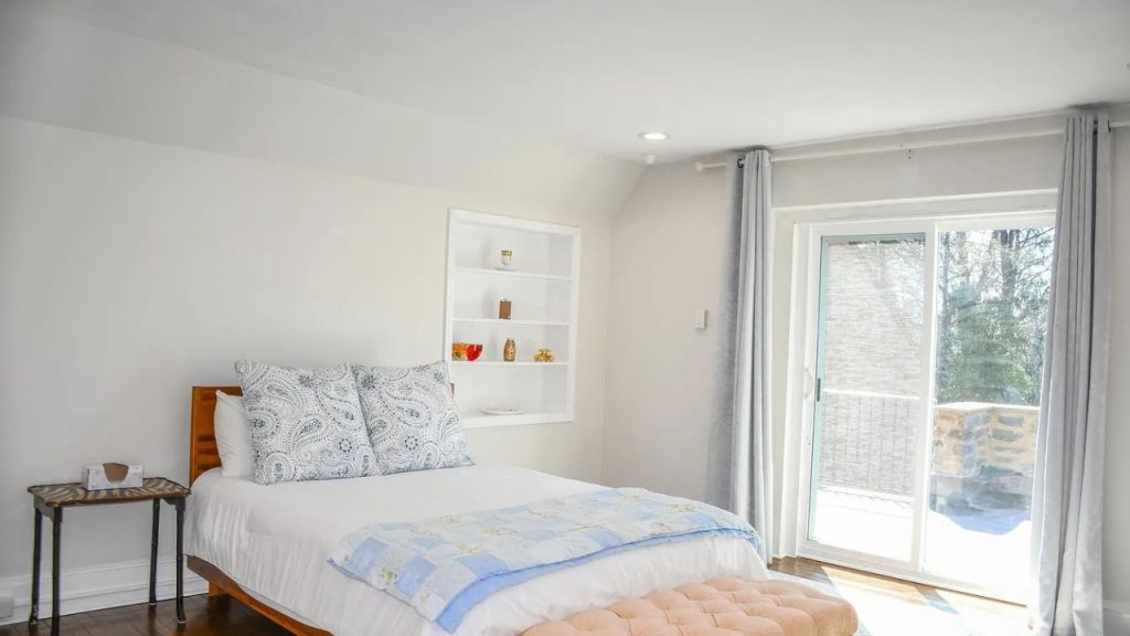 A serene bedroom with a cozy bed, sliding glass door, built-in shelf, and pink bench on hardwood floor.