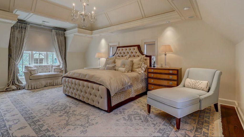 A serene bedroom with a tufted bed, chandelier, window seat, and chaise lounge on an ornate rug.
