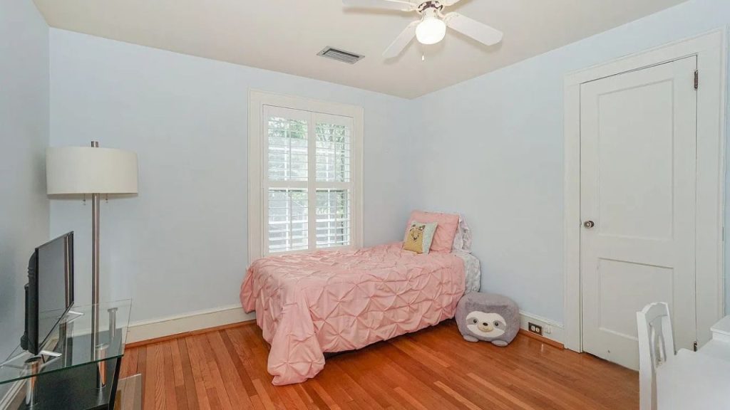 A playful kid’s room with a pink quilted bed, large window, ceiling fan, and a sloth plushie on hardwood floors.
