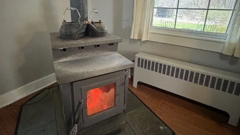 A cozy nook with a wood stove, vintage kettles, a large window, and a radiator on a hardwood floor.