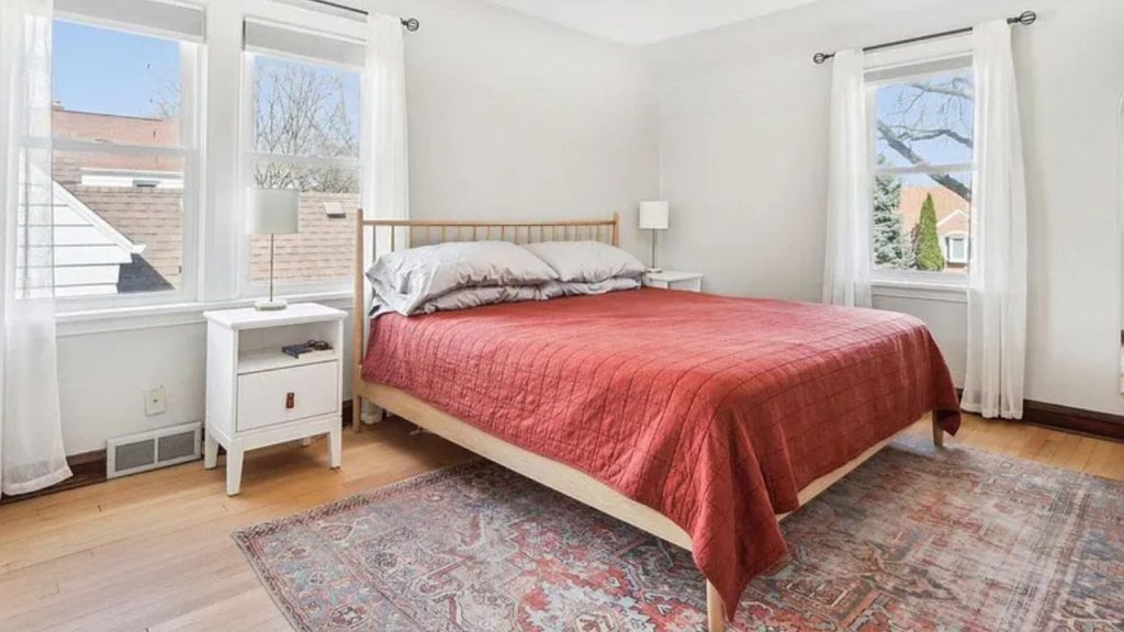 A cozy bedroom with a red quilted bed, white nightstand, and large windows.