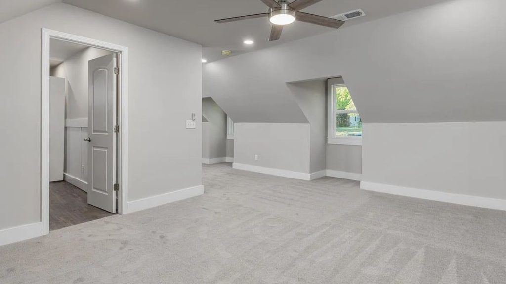 A spacious attic bedroom with slanted ceilings, a ceiling fan, large window, and soft carpeting.