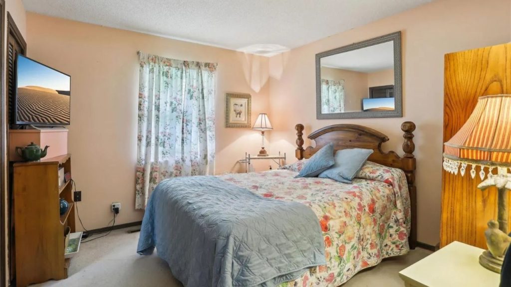 A peaceful bedroom with a wooden four-poster bed, floral quilt, mounted TV, and floral curtains, featuring a vintage lamp and mirror.