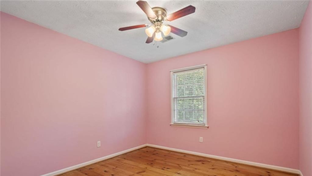 A cheerful bedroom with pink walls, wooden floors, a ceiling fan, and a large window with greenery.
