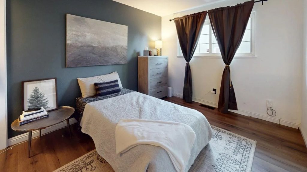 A cozy bedroom with a teal accent wall, white bed, wooden dresser, side table with books, and dark-curtained window.