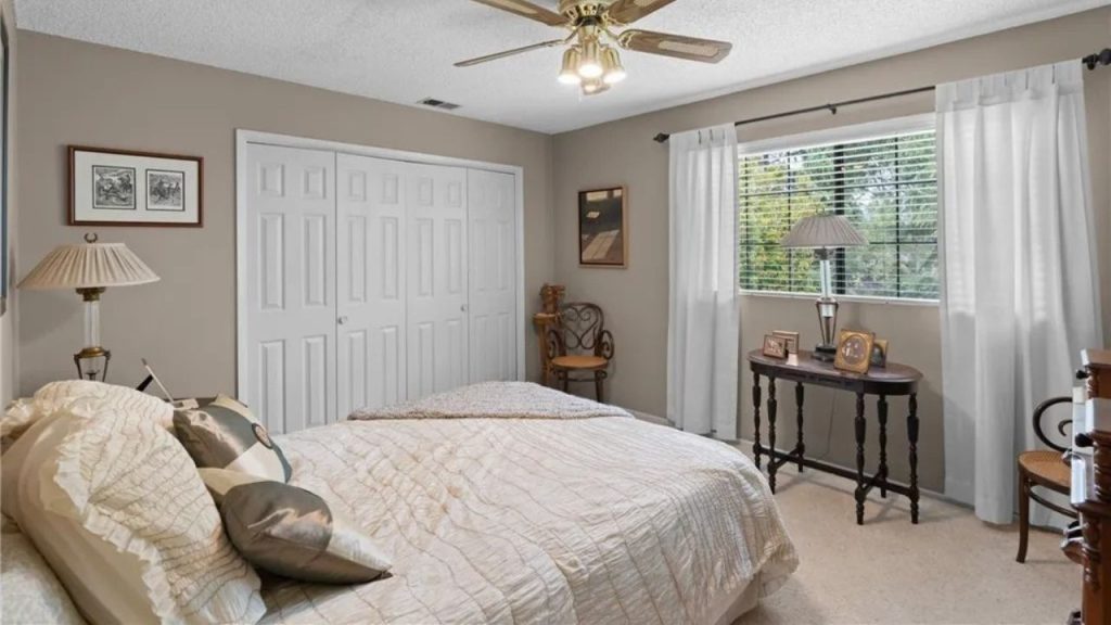 A warm guest bedroom with a cozy bed, wooden console, large windows, and a classic chair.