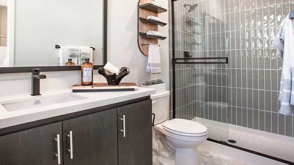 A minimalist bathroom with a dark vanity, white quartz countertop, gray subway tile walls, a glass-enclosed shower, a modern toilet, and a wooden shelf with towels.
