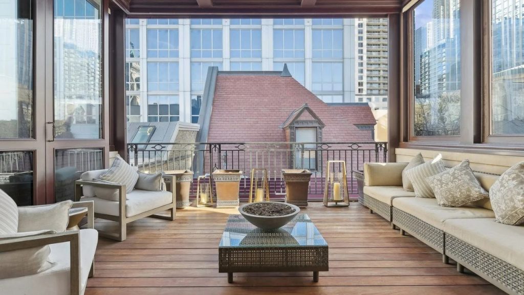 A cozy terrace with wicker furniture, plush cushions, a glass-top coffee table, and potted lanterns, set on a wooden deck with a city skyline view.