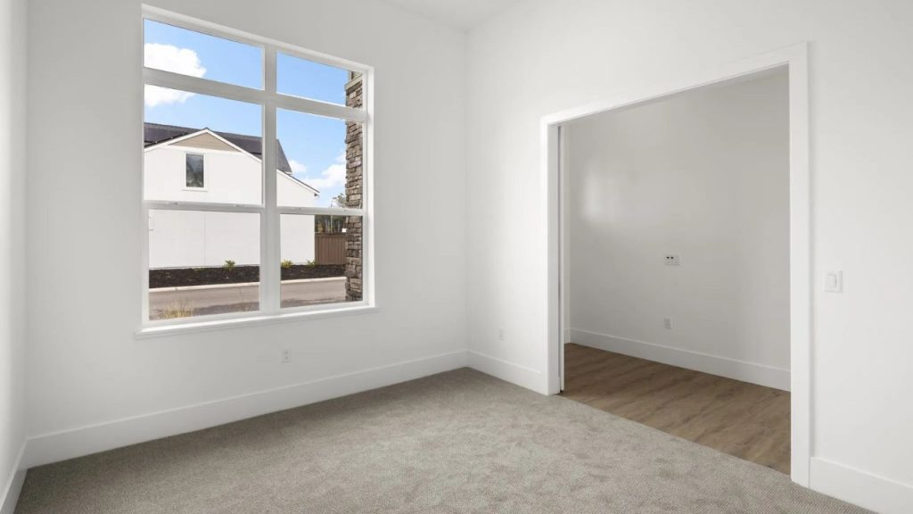 A bright primary suite with gray carpeting, white walls, a large grid-patterned window, and an open doorway to an ensuite.