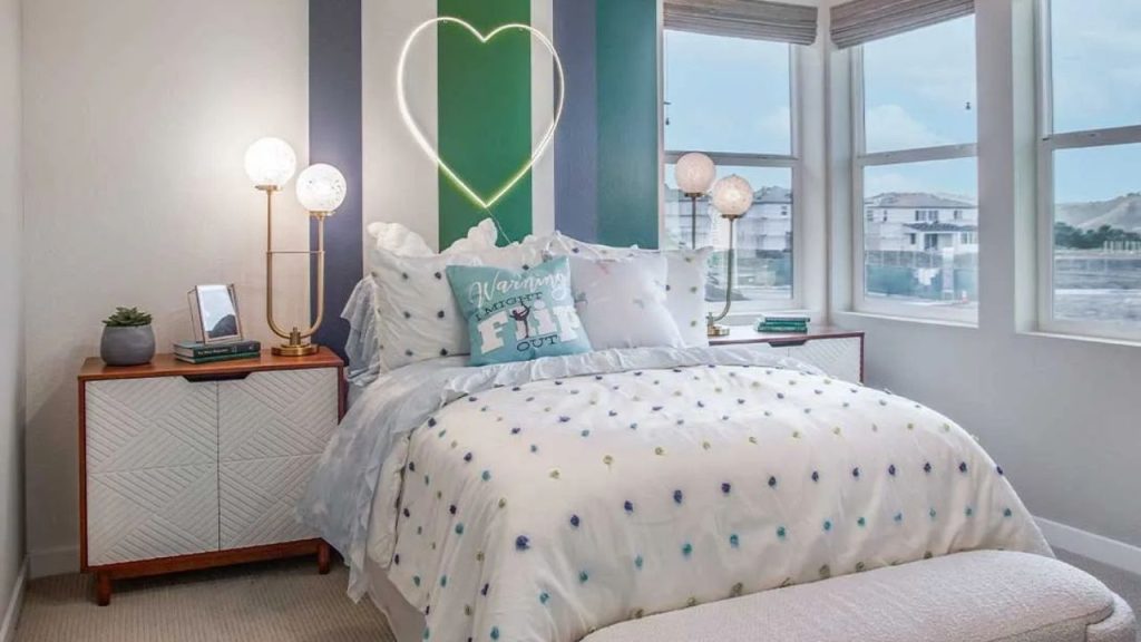 A bright bedroom with a white bed, blue polka-dot bedding, green accent wall, and a neon heart light.