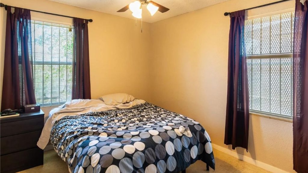 A cozy bedroom with a polka-dot bed, purple curtains, dark nightstand, and ceiling fan.