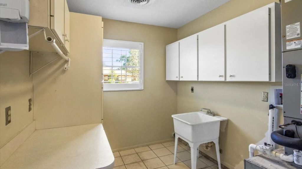 A laundry room with beige walls, tiled floors, a utility sink, white cabinets, a window with shutters, and a hanging rod for drying clothes.