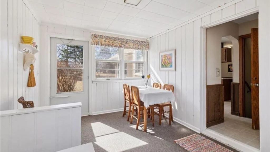Cozy sunlit breakfast nook with wooden table and chairs, surrounded by white paneled walls and large windows.