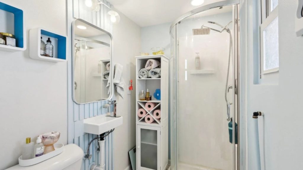Neatly designed small bathroom with shower, storage shelf, and soft blue accents.