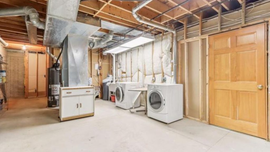 A basement laundry area with exposed beams, two white washers and dryers, a wooden door, and unfinished walls, exuding a practical charm.