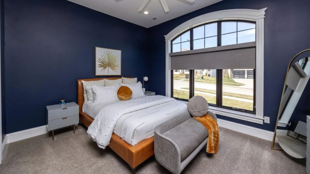 A vibrant bedroom with navy walls, a cozy bed, large arched window, and a stylish bench, radiating warmth and charm.