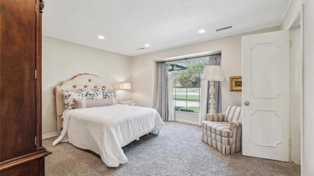 A tranquil bedroom with a white bed, striped armchair, large windows, and wooden furniture.