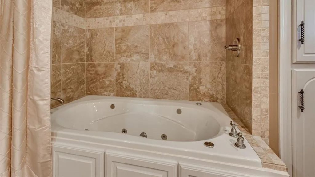 Cozy bathroom corner with a jetted tub, stone tiles, white cabinetry, and beige curtains.