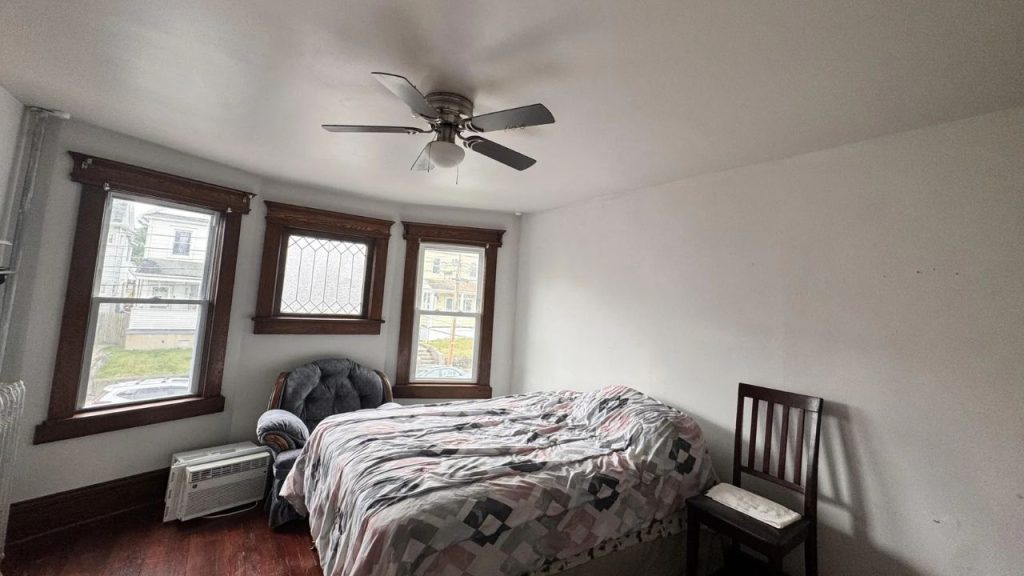 A cozy bedroom with wooden floors, a patterned bed, armchair, and large windows.
