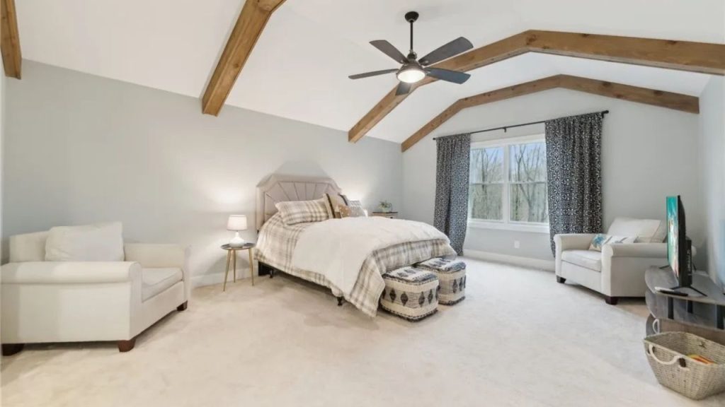 A cozy bedroom with wooden beams, a plush bed, and armchairs near large windows.