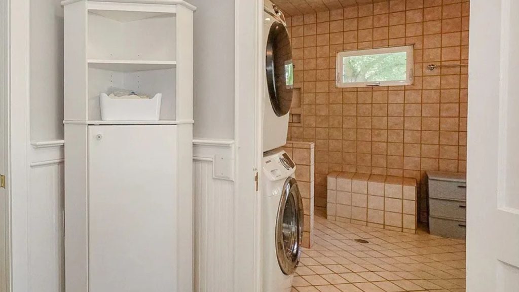 A functional laundry nook with a stackable washer-dryer, corner shelves, and a tiled shower room with a small window.