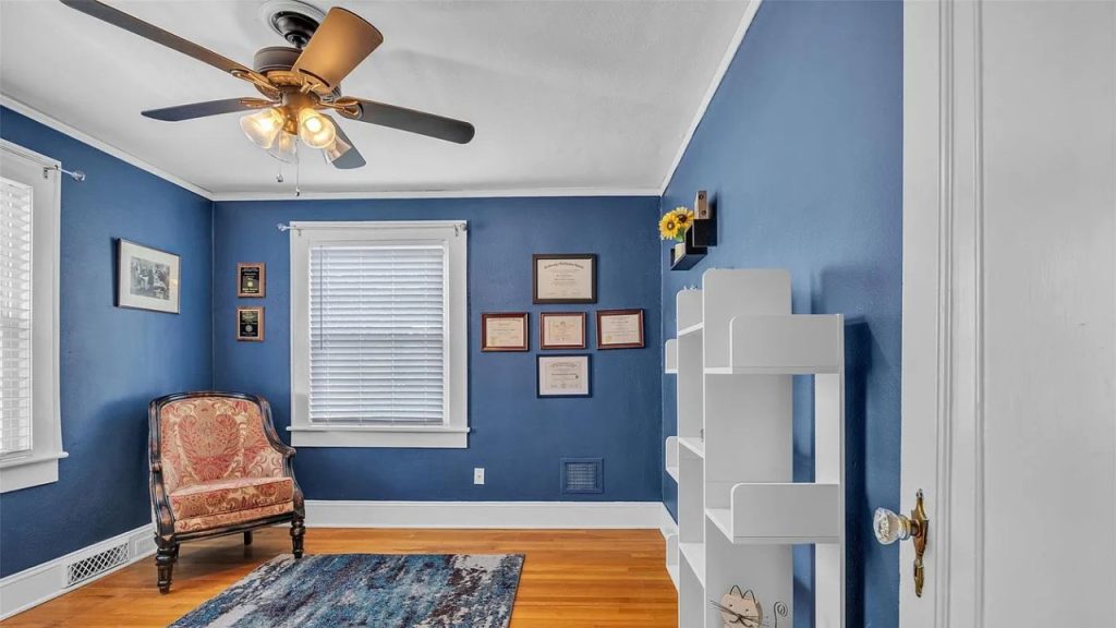 A quiet study corner with blue walls, a vintage armchair, white shelves, and a colorful rug under a ceiling fan.