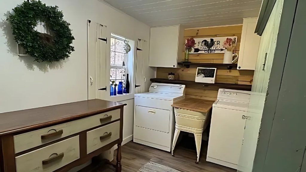 A rustic laundry room with white washers, wooden dresser, and a window with sheer curtains.