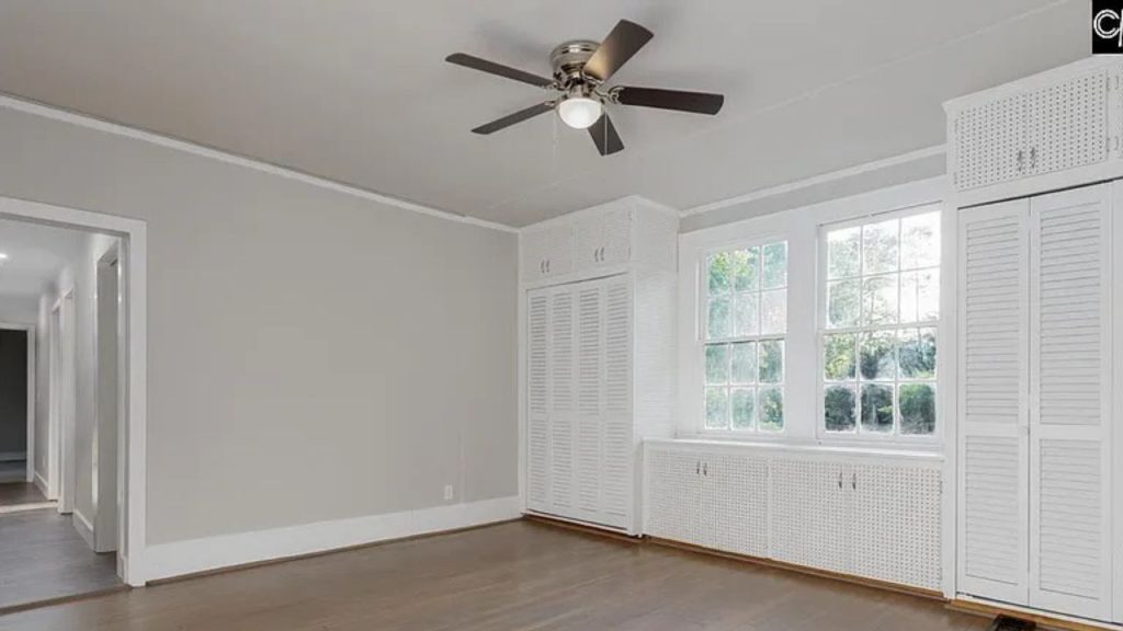 A bright bedroom with large windows, a ceiling fan, built-in wardrobes, and hardwood floors.