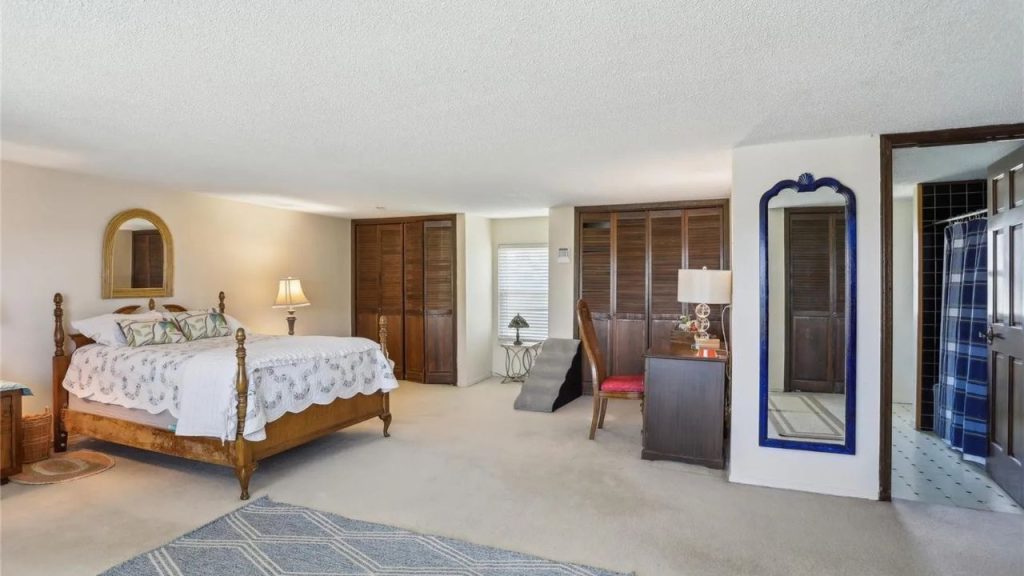 A tranquil bedroom with a wooden four-poster bed, louvered closets, blue-framed mirror, and geometric rug under soft natural light.
