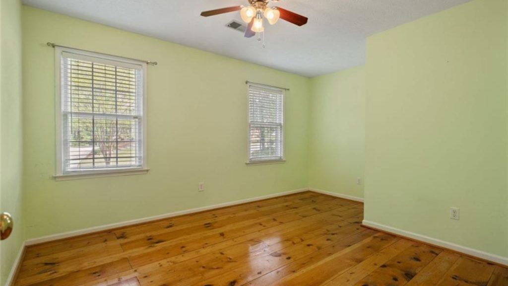 A bright bedroom with light green walls, wooden floors, two windows, and a ceiling fan.