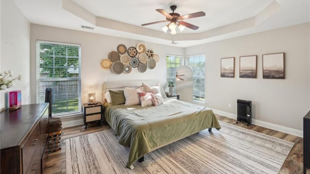 A peaceful bedroom with a green bed, woven wall art, large windows, and a ceiling fan.